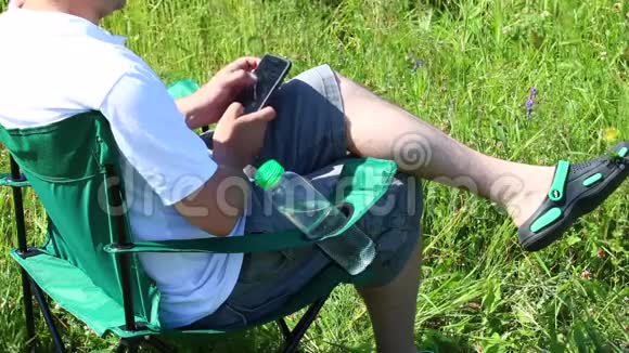 一个人坐在一片灯火通明的草地上的野餐椅上他在休息在做智能手机草和花从夏天开始摇摆视频的预览图