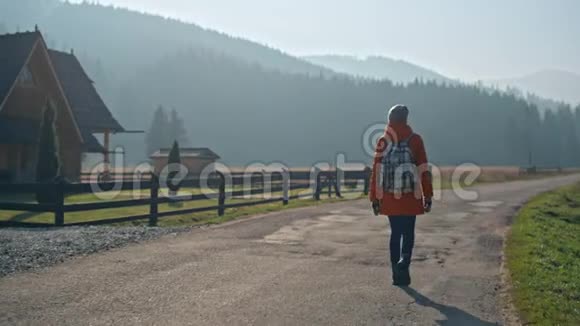 带背包的女孩走在乡村道路上在秋天的房子和山脉附近视频的预览图