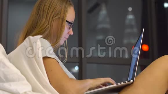 一个带眼镜的有趣的年轻女人她用笔记本电脑坐在床上笑着她在打字视频的预览图