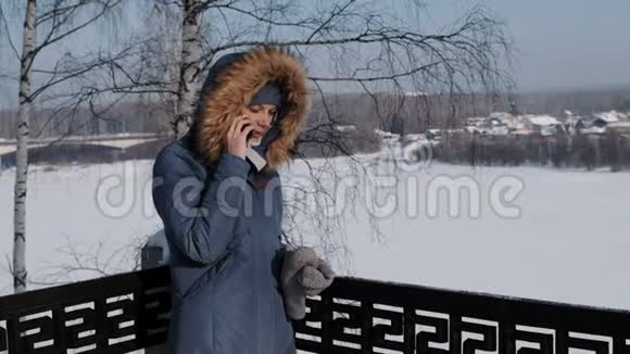穿着蓝色羽绒服的女人在冬季公园打电话给她的手机视频的预览图