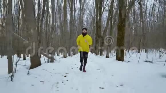 穿着黄色外套的蓄胡子运动员在冬季森林中奔跑的手持镜头视频的预览图
