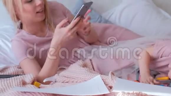 年轻迷人的金发妈妈和她穿着粉红色连衣裙的小可爱女儿在手机上拍了一张有趣的照片视频的预览图