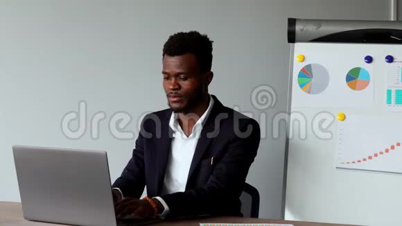 美国黑人男子坐在笔记本电脑前穿着西装穿着白色衬衫坐在图表和桌子的背景上视频的预览图