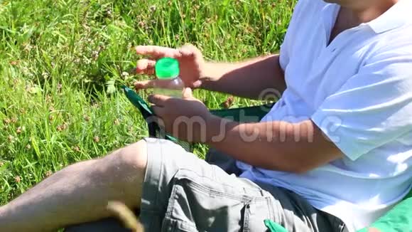 一个人坐在一片灯火通明的草地上的野餐椅上他休息关上一瓶水然后换腿草和花会摇摆视频的预览图