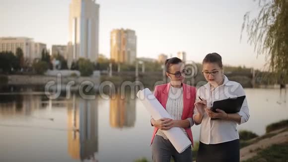 两个女人手里拿着蓝图在城市的海滨背景是高层建筑讨论着一个视频的预览图