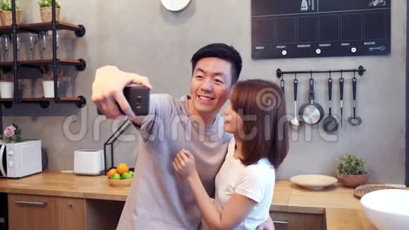 快乐的亚洲年轻夫妇在家厨房做饭时用智能手机自拍视频的预览图