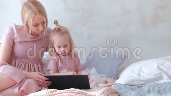 年轻迷人的金发女人和她的小迷人的女儿穿着粉红色的裙子静静地看着平板电脑里的东西视频的预览图