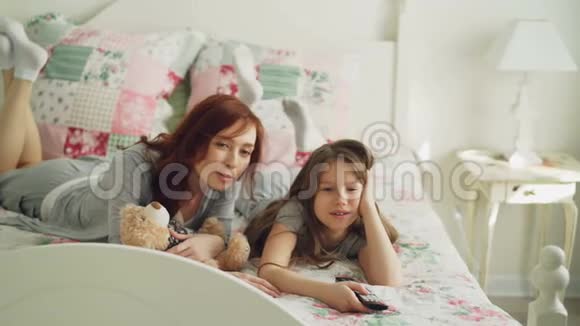 美丽快乐的母亲和小女儿在电视上看卡通电影在家里躺在床上大笑视频的预览图