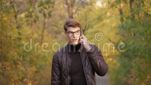 阳光明媚的一天一个年轻有魅力的男人打着电话在秋天公园散步视频的预览图