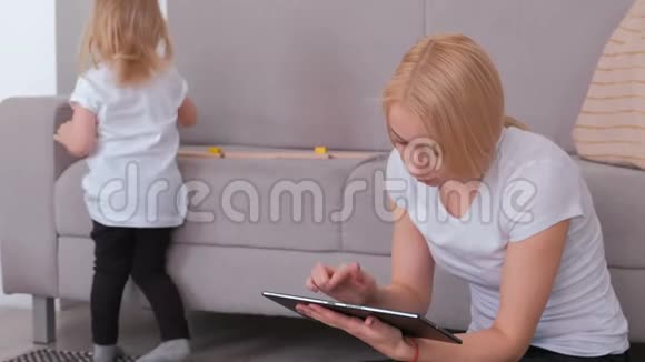 年轻迷人的金发妈妈在看平板电脑而她迷人的女儿在玩木块视频的预览图