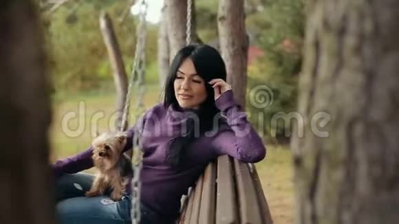 带着狗在长凳上荡秋千的女孩那个迷人的年轻女孩和狗坐在公园的长凳上慢动作视频的预览图