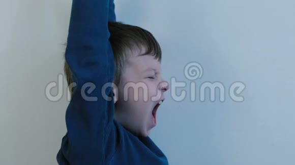 七岁的男孩穿着一件蓝色衬衫在浅色背景下打着哈欠双手向上伸展视频的预览图