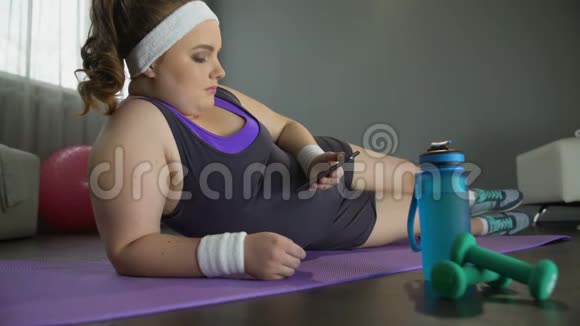 肥胖的懒女人躺在地上手里拿着智能手机而不是训练视频的预览图