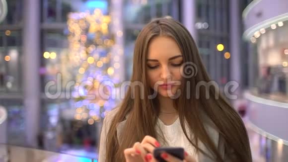 美丽的快乐女孩长发在智能手机上写着一条信息站在商场里慢动作妇女使用视频的预览图