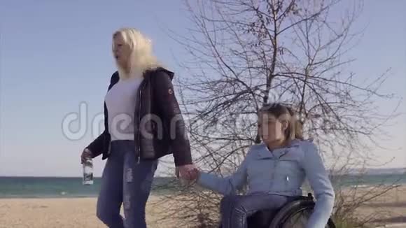 家庭休闲坐轮椅的年轻残疾妇女和她的母亲在海边散步说话玩得很开心视频的预览图