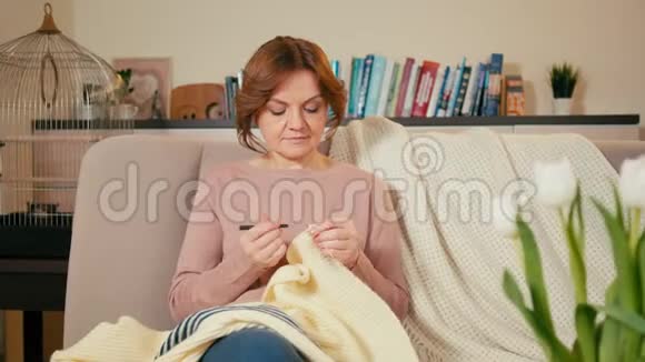 漂亮的成年女人正在家里舒适的房间里编织毯子坐在沙发上视频的预览图