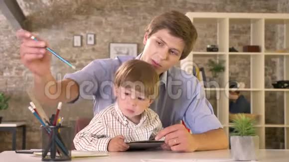 年轻的父亲抱着他的小儿子写着纸孩子拿着平板电脑坐在现代的办公室里视频的预览图