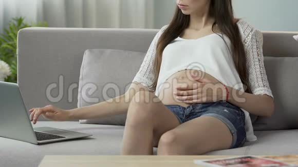 期待女人坐在沙发上用笔记本电脑选择分娩中心视频的预览图