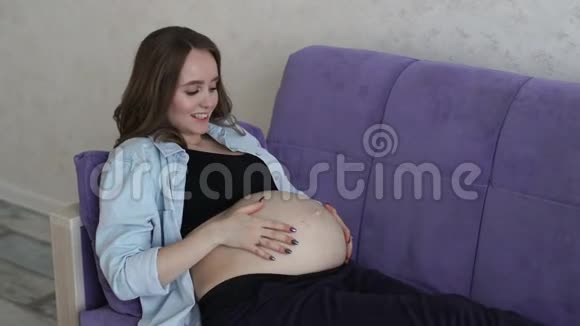 一位快乐的孕妇坐在家里的沙发上用手抚摸肚子同时与婴儿交流视频的预览图