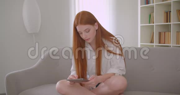 一位年轻漂亮红发女郎在舒适的公寓里坐在沙发上浏览平板电脑的特写肖像视频的预览图