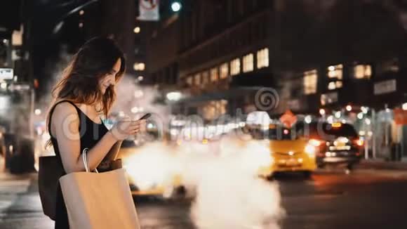 年轻漂亮的女人带着购物袋站在纽约市中心的交通中使用智能手机视频的预览图