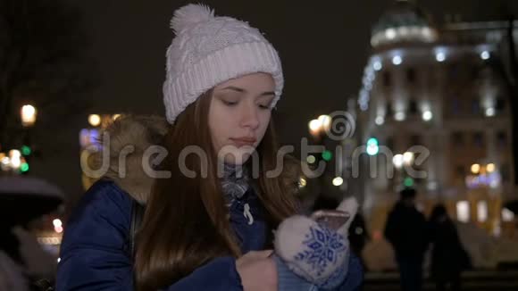 冬天一个戴白帽子的女孩走在街上用智能手机看一条信息视频的预览图