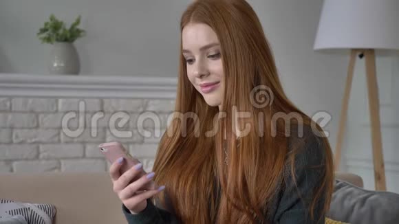 年轻漂亮的红发少女使用智能手机微笑发短信滚动滑动惊讶的情绪回家视频的预览图