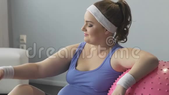 超重女孩坐在地板上靠在健身球上屈臂满意视频的预览图