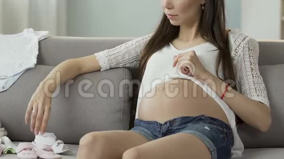 怀孕的女性坐在沙发上拿着婴儿鞋看着它们婴儿时尚视频的预览图
