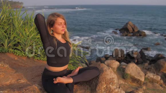 穿着运动服的女士坐在悬崖上在海边做瑜伽姿势视频的预览图