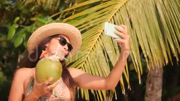 在热带地区用智能手机和饮用椰子鸡尾酒制作自拍照的年轻混血女游客视频的预览图