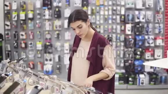 年轻美丽的黑发女孩微笑滚动一个iPhone绑在展示货架在电器商店选择新的视频的预览图
