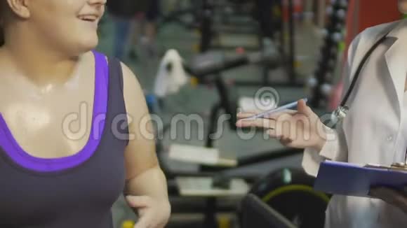 肥胖女孩在健身房跑步机上锻炼和护士交谈看体重视频的预览图