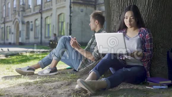 年轻女人用笔记本电脑男人用笔记本做笔记坐在树下视频的预览图