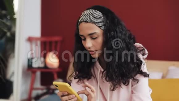 美丽的非裔美国女孩聊天和短信智能手机坐在床上在家里年轻女子视频的预览图