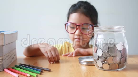 亚洲小女孩把硬币放在一个透明的玻璃罐子里的桌子上比喻省钱的概念声音选择关注罐子视频的预览图