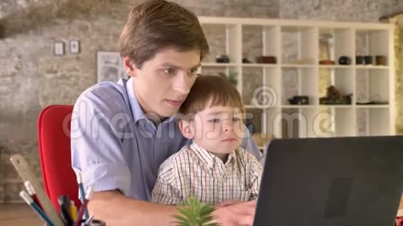 年轻的父亲抱着他的小儿子用笔记本电脑工作忙着坐在现代的办公室里视频的预览图