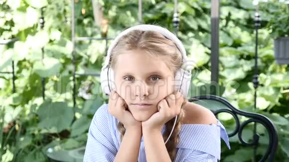 一幅漂亮女孩坐在街头咖啡馆的肖像桌子上有笔记本头上有耳机听音乐视频的预览图