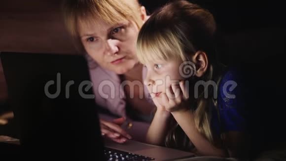 年轻的母亲和女儿一起在笔记本电脑上玩躺在床上屏幕上的光视频的预览图