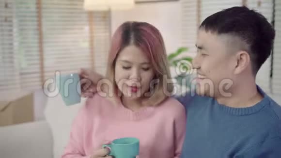 迷人的亚洲甜蜜夫妇享受爱情时刻在客厅的沙发上喝着温暖的咖啡或茶视频的预览图