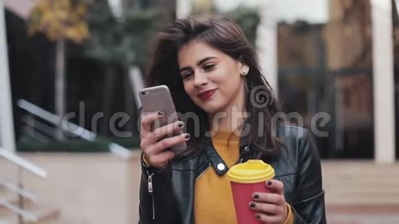 年轻快乐的女人使用智能手机喝咖啡去户外沿着城市的街道走慢动作视频的预览图