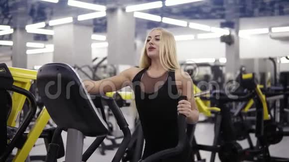 年轻迷人的金发女郎微笑着在健身馆穿着黑色运动服的椭圆教练模拟器上蹬蹬视频的预览图