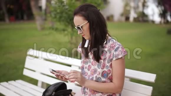 漂亮的女人坐在公园的长椅上用智能手机视频的预览图