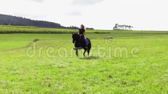 美丽的金发女郎骑着一匹黑马骑着田野视频的预览图