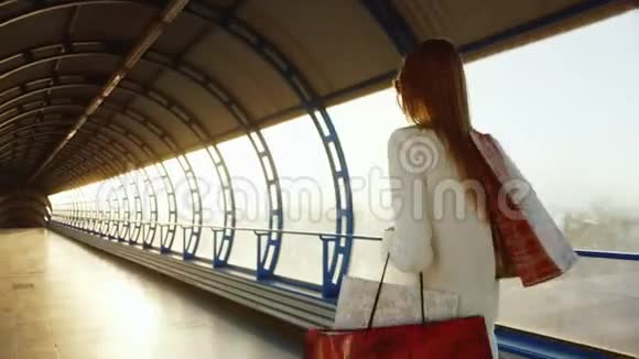 后景带购物袋的快乐女人微笑沿着现代化的玻璃隧道行走持有智能手机视频的预览图