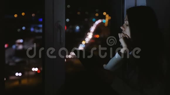 在黑暗的夜晚女人坐在窗台上看着街道使用平板电脑上网视频的预览图