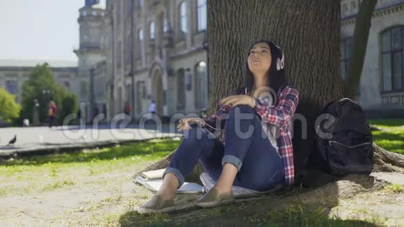 戴着耳机欣赏音乐的亚洲女性坐在树下视频的预览图