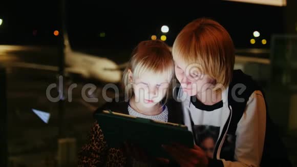 妈妈和女儿在等航班一起在平板电脑上玩延误或待定飞行概念视频的预览图