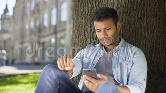 年轻男性坐在树下用手机阅读信息令人沮丧的新闻视频的预览图