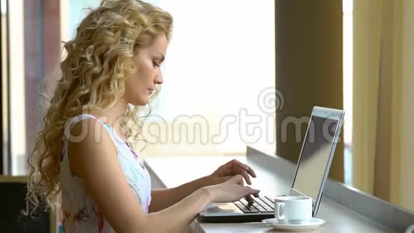 漂亮的金发女孩在咖啡馆里用笔记本电脑穿衣服年轻的女人喝咖啡在笔记本上工作视频的预览图